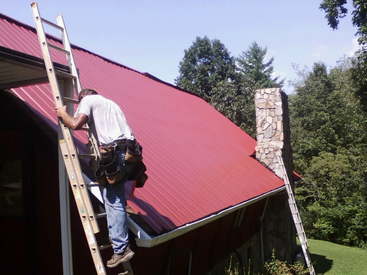 Roofer on a ladder beside a red metal roof and stone chimney — Soffit & Fascia Repair by Roofing Brothers in Girdletree